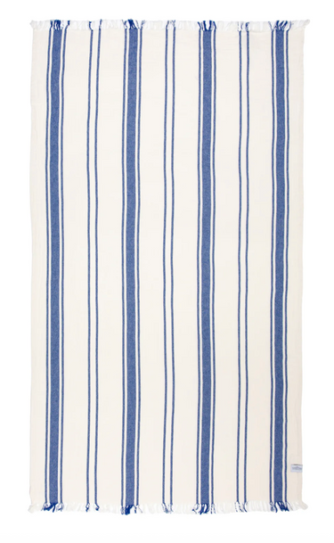A rectangular beach towel with a white background and evenly spaced vertical stripes in two shades of blue, alternating between a thinner and a thicker stripe. Both the top and bottom edges of the towel have white fringe with hints of blue. A small label is visible on the bottom right corner.